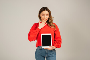 surprised woman in wireless earphones holding digital tablet with blank screen isolated on grey