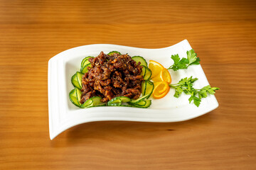 Chinese popular recipe of caramelized beef strips with sesame seeds on raw cucumber slices and orange slices with parsley on a white tray