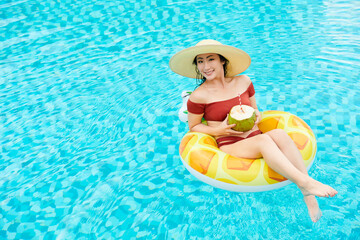 Portrait of cheerful young Asian woman in straw hat sitting with coconut cocktail on inflatable ring in swimming pool