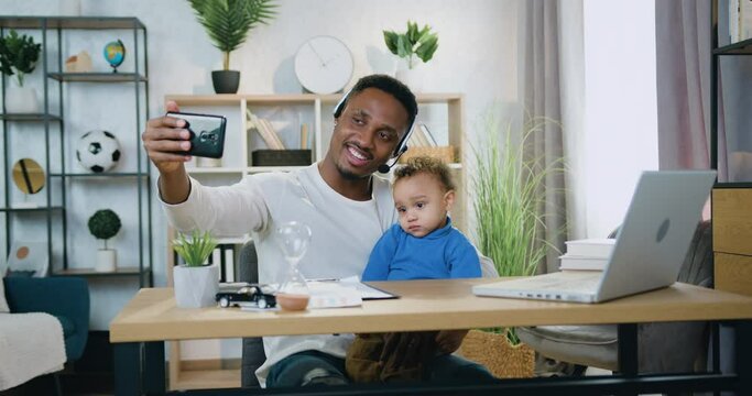 Good-looking smiling happy modern black-skinned father in headset making selfie together with his small son at home