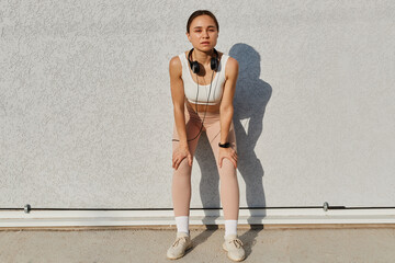 Naklejka premium Full length portrait of sporty slim woman wearing white top and beige leggins, having headphones over neck, leaning to wall outdoor, posing after training.