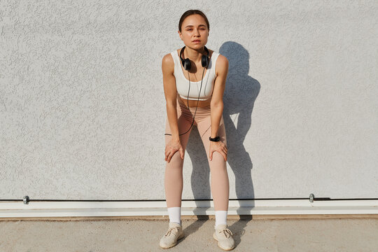 Full length portrait of sporty slim woman wearing white top and beige leggins, having headphones over neck, leaning to wall outdoor, posing after training.