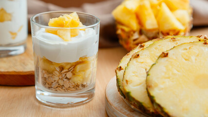 Delicious pineapple dessert. Breakfast dessert with oat granola, greek yogurt and pineapple in layers in glass with ingredients chopped fresh juicy pineapple on wooden table. Long web banner
