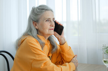 Beautiful old grandmother with grey hair and face with wrinkles is using smartphone, talking with someone and sitting at the table at home on window background, mother's day, happy retirement