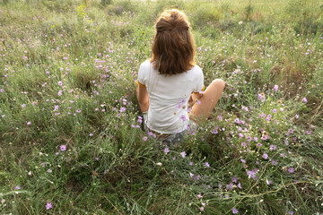 Fototapeta premium Woman sitting on the green grass and among wild flowers.Sunset time on the meadow
