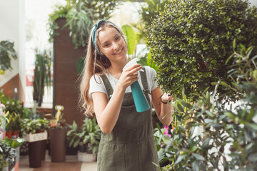 Young female gardener in spraying myrtle tree houseplant in flower store or greenhouse