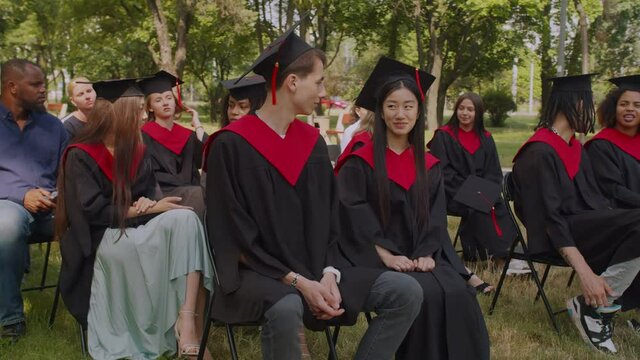 Positive interracial diverse college friends in academic gowns and mortarboards talking and discussing, sitting on chairs on green lawn, preparing for graduation ceremony at university campus.