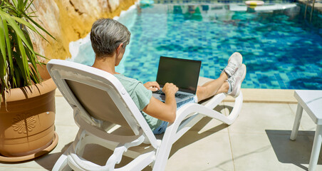 Rear view man relaxing on lounge by the swimming pool at resort hotel, using laptop computer, typing text on keyboard, work and travel concept