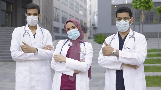 Team of three young Arabian male and female doctors in white coats and protective masks, standing outside modern clinic building with arms crossed and showing thumbs up