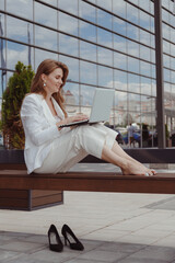 Barefoot businesswoman in white suit sitting on bench with laptop.