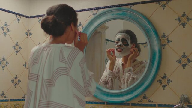 Young adult woman applying facial cosmetics mask looking like panda. Mirror reflection of woman wearing panda mask.