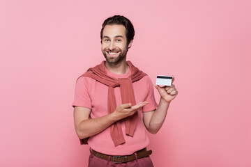 Positive man pointing at credit card while standing isolated on pink