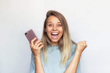 A young pretty caucasian impressed excited smiling blonde woman in a grey t-shirt with a mobile phone in her hand is happy about the news or lottery win isolated on a light color gray background