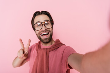 Happy man in eyeglasses showing peace sign isolated on pink
