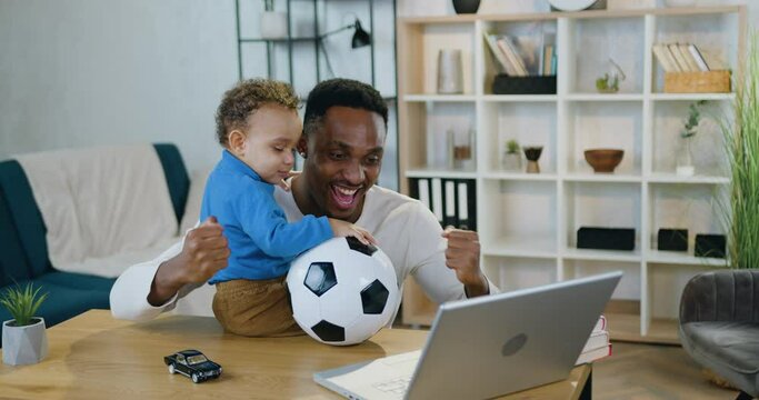 Attractive cheerful happy caring young african american dad-football fan rejoycing from victory of favourite team and hugging his cute small son,child care and leisure concept