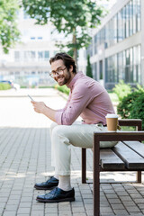 Smiling businessman using smartphone near coffee to go on bench