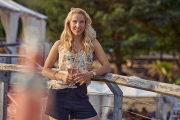 Pretty relaxed blond woman enjoying a tropical cocktail on summer vacation standing leaning on wooden railings looking at the camera with a friendly smile and copyspace