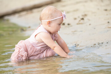 A beautiful little girl sits on the river bank and plays with sand and water. Child and nature.