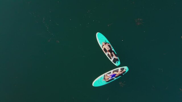 Surfers couple lying on surfing board, man and woman swimmers relaxing on blue surfboards top view. Professional drone shooting of people on sup boards in lake. Nature and calmness.