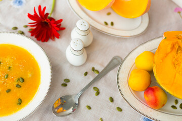 Plate with colorful pumpkin soup, pumpkin cut into pieces on porcelain plates, pepper shaker, salt shaker, spoon, red gerbera flower and green pumpkin seeds  on white tablecloth  