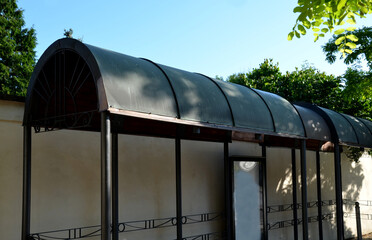 metal bus stop by the beige plaster wall. the roof is made of sheet metal bent into an arch....