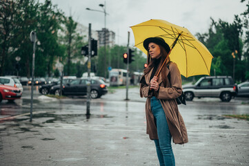 Woman standing near the crossroad during rain and holding a yellow umbrella