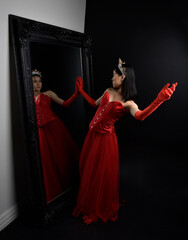 Full length  portrait of beautiful young asian woman wearing red corset, long opera gloves and ornate crown headdress. Graceful posing against a full length mirror with a dark studio background.