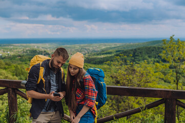 Couple of hikers using using smartphone for orientation while standing on the lookout - obrazy, fototapety, plakaty