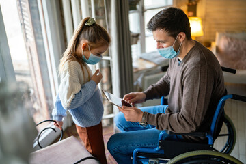 Father with disability in wheelchair using tablet at home with child while wearing masks.