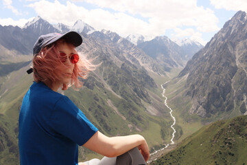 A young woman traveler in a baseball cap and sunglasses with pink hair sits on a cliff in the mountains, admiring the views of the wildlife. Hiking on the Tien Shan mountain range, Kyrgyzstan