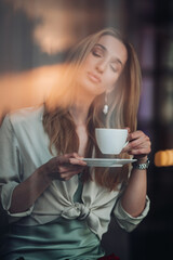 Modern romantic beautiful young girl enjoying coffee cup fragrance with closed eyes while relaxing in cafe indoors