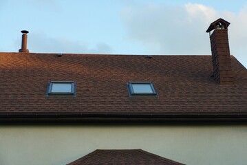brown tiled roof of a gray private house with a two windows and one brick chimney against a blue sky