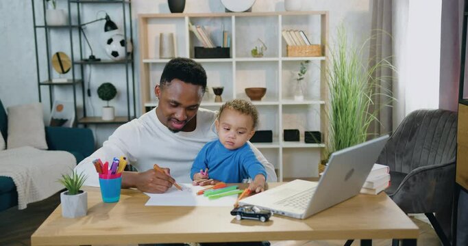 Child care concept where good-looking happy relaxed caring young black-skinned man drawing together with his cute baby boy with coloured felt-tip pens at home,front view