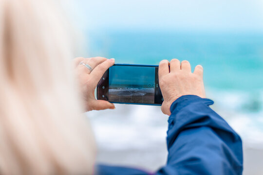 Tourist caucasian blonde woman smiles, takes pictures on the phone on the beach near the ocean.