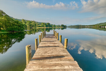 A wooden jetty on Coniston Water in the Lake District, shot in landscape, looking straight down the jetty to the hills beyond, with clouds reflected in the water