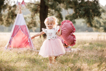Girl are dressed in pink dress and holding big pink heart in hands. Mom, dad are sitting next to wigwam decoration in the park. Family spending time outdoor in summer, having fun together. © Andriy Medvediuk