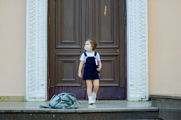 a beautiful little girl, a schoolgirl, stands outside the school during the day, a medical mask on her face