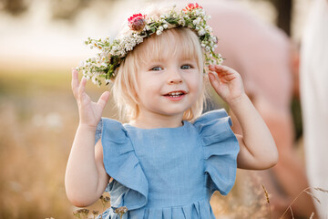 little girl is wearing a flower wreath on her head in a field on summer sunny day. baby in a blue...