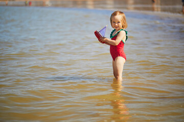 Adorable toddler girl in red swimsuit playing with wooden toy boat in water on the beach in...