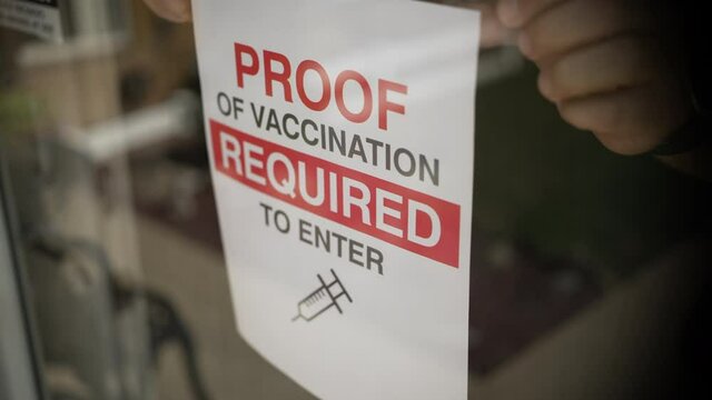 A small business owner places a VACCINATION REQUIRED sign on his front door window. Some businesses started to customers to show proof of their coronavirus vaccination before entering.  	