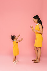 full length of asian mother looking at toddler daughter pointing at soap bubble on pink