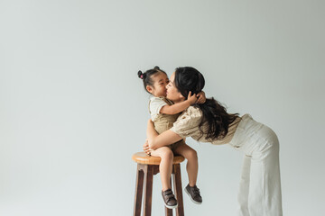 asian toddler girl sitting on chair and kissing mother isolated on grey