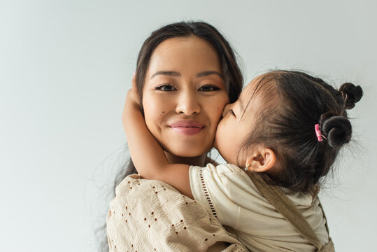 asian toddler girl kissing cheek of happy mother isolated on grey
