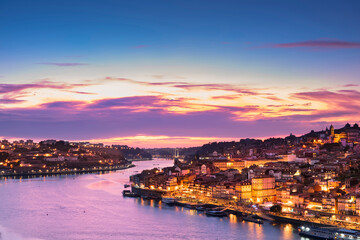 View from the Luis I Bridge of the Douro embankment at sunset, the city lit by lights, boats and houses along the coast.
