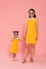 cheerful asian woman holding hands with toddler daughter in sunglasses on pink