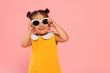 cheerful asian toddler kid in yellow dress adjusting sunglasses isolated on pink