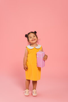 Full Length Of Pleased Asian Toddler Kid In Yellow Dress Holding Present On Pink