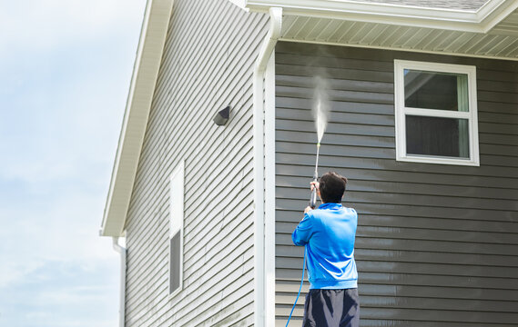 Man in blue jacket clean dusk and dirt from exterior siding and under roof by high-pressure nozzle spray with water soap cleaner, weekend chore on sunny day. Cleansing and maintenance service concept.