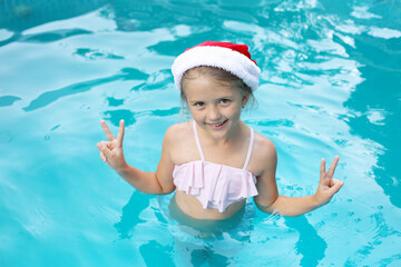 a beautiful happy girl in a Santa Claus hat in a pink swimsuit is standing in the pool for the new year