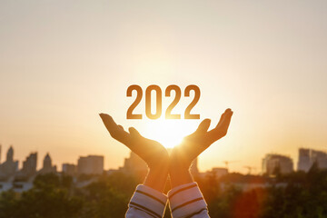 2022 is supported by hands on the background of a sunny sunset. - obrazy, fototapety, plakaty